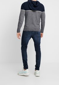 Granatowo-szara dzianinowa bluza z kapturem o teksturowanym korpusie, z prążkowanymi rękawami, kontrastującym poziomym paskiem, zestawiona z ciemnymi obcisłymi dżinsami i trampkami.