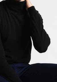 Czarny sweter z wysokim kołnierzem, z ribbowanym wykończeniem i długimi rękawami, wykonany z miękkiego materiału. Noszony z ciemnoniebieskimi spodniami.