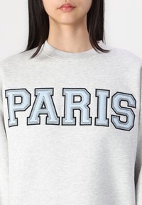 Felpa grigia con la scritta "PARIGI" in lettere grasse e contornate, arricchita da accenti azzurri. Il materiale sembra morbido e presenta un classico collo a giro.