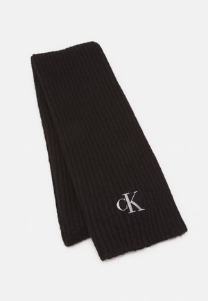 Écharpe noire en maille côtelée pliée avec logo blanc "cK" brodé près d'une extrémité sur un fond blanc.