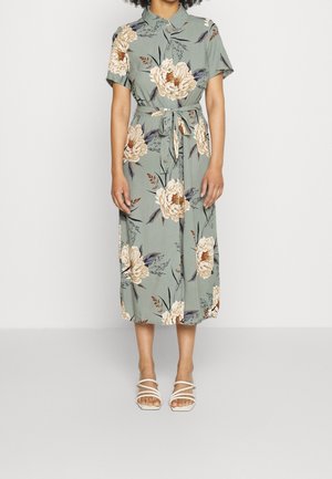 Kvinde iført en grøn midi-kjole med blomsterprint, korte ærmer, krave, bånd i taljen og hvide sandaletter med stropper.