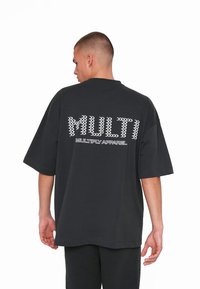 Sort oversized t-shirt med fed, hvid tekst "MULTI" og "MULTIPLY APPAREL" i forskellige stilarter på ryggen, lavet af bomuldsmateriale.