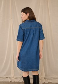 Hosszú barna hajú nő kék farmerruhában és fekete térdig érő csizmában, bézs színű textilminta előtt áll.