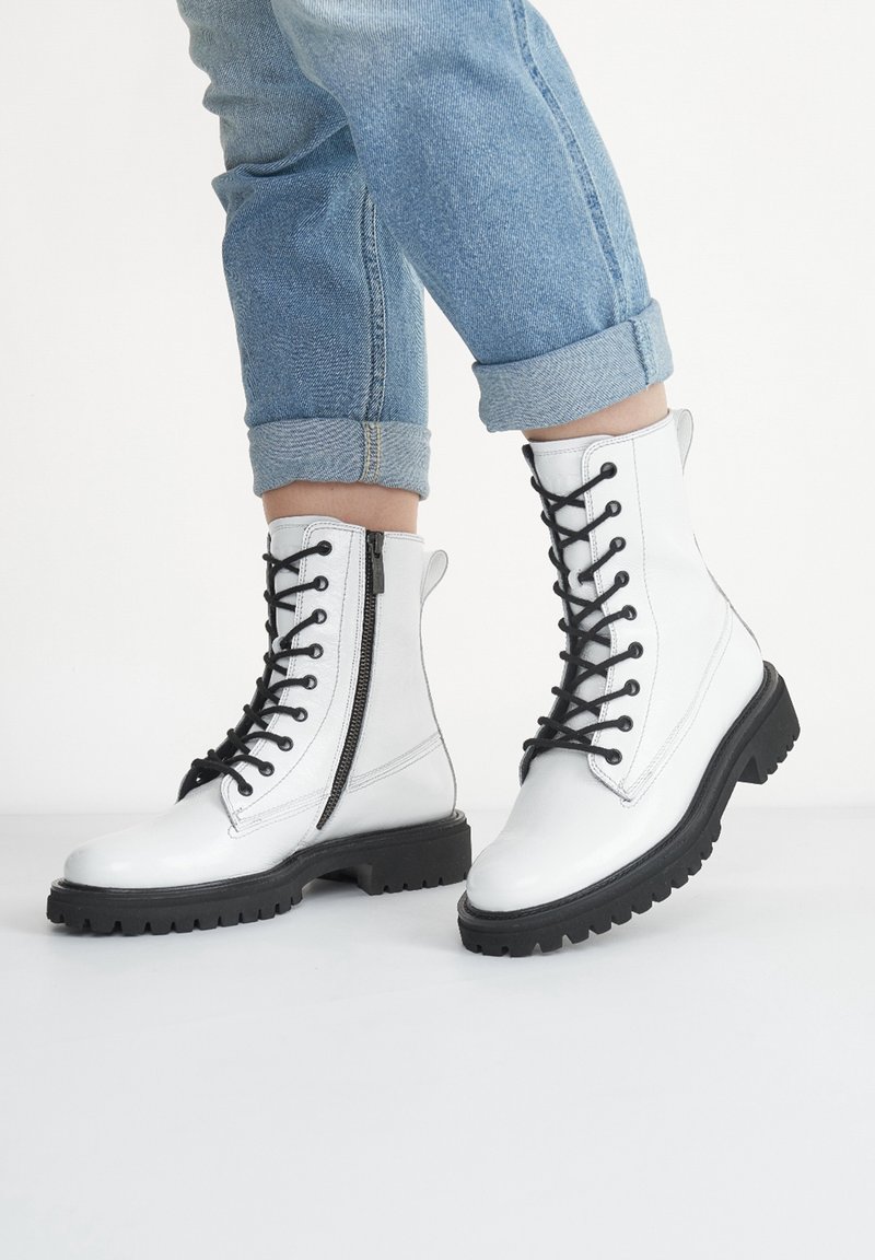 Bottes de combat en cuir blanc avec une semelle en caoutchouc noire, dotées d'une fermeture éclair frontale et d'un design à lacets. Finition texturée, hauteur à la cheville avec une forme robuste.