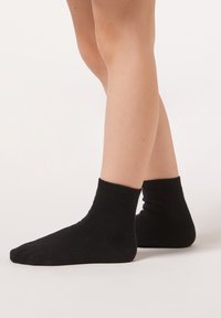 Svarta ankelsockor med ribbad kant och slät yta, visade på fötter med ljus hudton mot en neutral bakgrund.