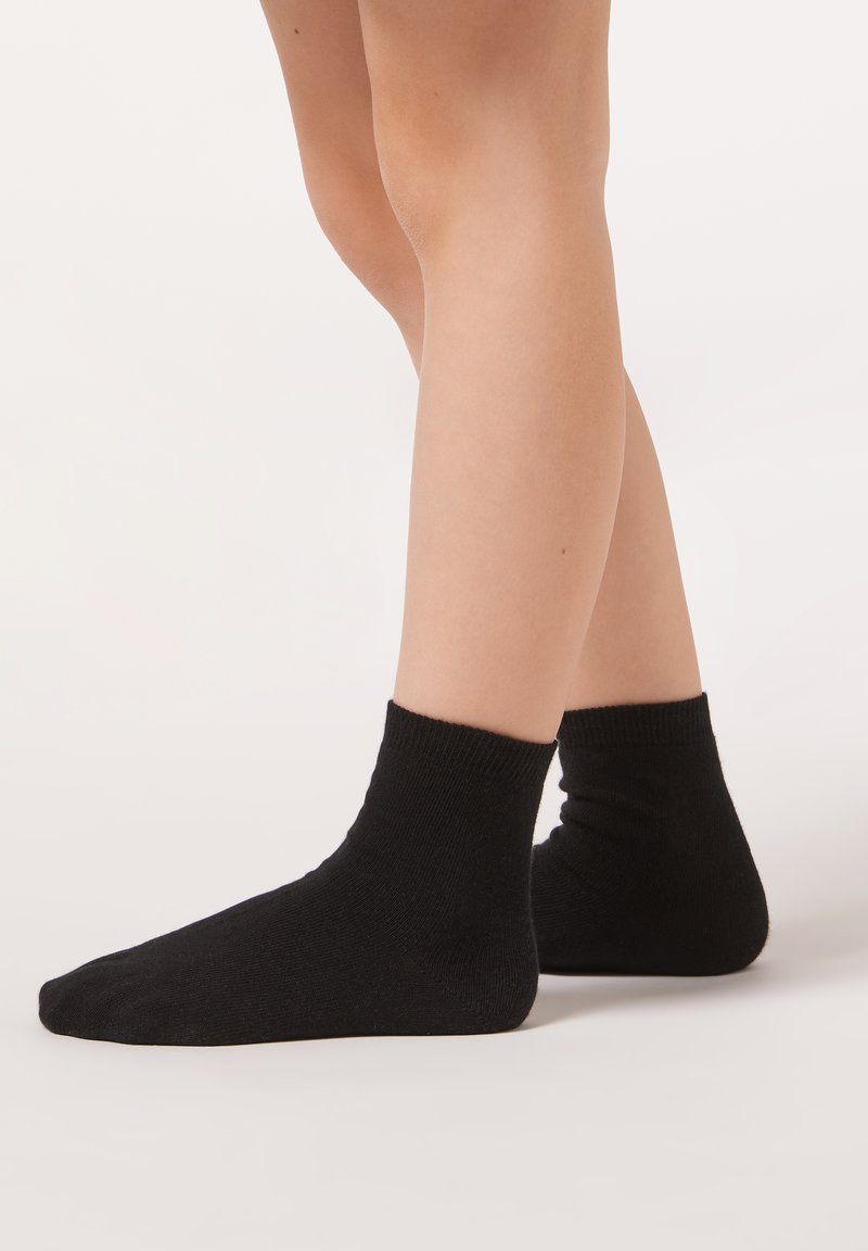 Svarta ankelsockor med ribbad kant och slät yta, visade på fötter med ljus hudton mot en neutral bakgrund.