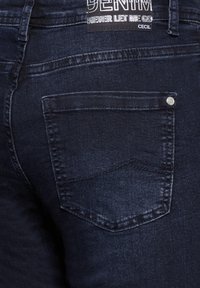 Jean en denim bleu foncé avec une poche arrière dotée d'une rivet, des coutures soignées et une étiquette portant la mention "DENIM NE ME LAISSE JAMAIS PARTIR CECIL."