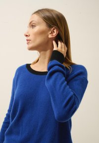 Modrý vlněný svetr s černým výstřihem a manžetami, který má volný střih a žebrované detaily na rukávech. Hladká textura.