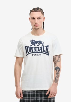 Ung mand med flettet hår iført hvid Lonsdale London T-shirt og sorte ternede bukser, der viser tattooet højre underarm på ensfarvet baggrund.