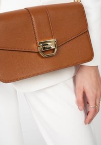 Braune Lederhandtasche mit einem strukturierten Design, ausgestattet mit einem goldenen Verschluss und einer strukturierten Oberfläche, gehalten von einer Person in Weiß gekleidet.