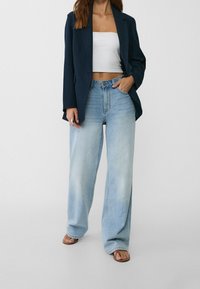 Kvinna som bär en marinblå kavaj, vit tube-top, ljusblå vida jeans och sandaler, stående mot en slät vit bakgrund.