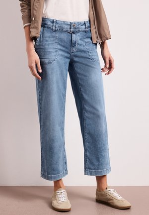 Lichtblauwe cropped wide-leg jeans gecombineerd met beige sneakers en een beige jas over een witte top, staand tegen een effen achtergrond.