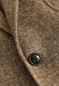 Bruine tweed stof met een fijne textuur en vlekken van donkerdere tinten. Bevat een enkele zwarte knop met vier gaten genaaid aan de linkerkant.