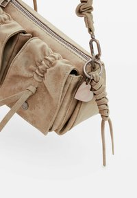 Sac à main en daim beige avec détails froncés, fermeture éclair, bandoulière tressée et un pendentif en forme de cœur. Présente une texture lisse.
