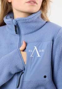 Giacca in fleece azzurro chiaro con colletto alto, tasca con zip e logo ricamato. Texture morbida e elementi di design minimi.