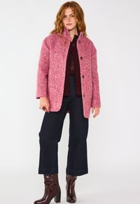 Cappotto in lana rosa, strutturato, con una vestibilità su misura e chiusura a bottoni. Abbinato a pantaloni flare in denim scuro e stivali in pelle marrone con tacco.