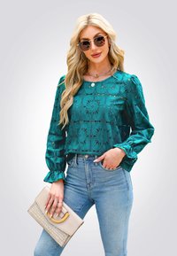Blusa in pizzo color turchese con motivo floreale, maniche a sbuffo e scollatura rotonda, abbinata a jeans azzurri a vita alta e una clutch beige.