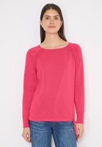 Femme aux longs cheveux noirs portant un pull à manches longues rose vif et un jean bleu, debout devant un fond blanc uni.