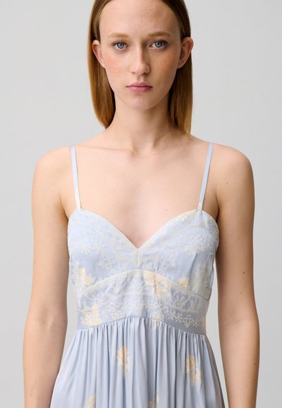 Giovane donna con capelli castano chiaro lisci che indossa un vestito azzurro pallido con sottili spalline e delicati ricami floreali, in piedi davanti a uno sfondo semplice.