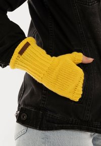 Gehaakte gele vingerloze handschoen met ribbelstructuur en manchet, met een bruine label. Draag op een zwarte jas tegen een lichte achtergrond.