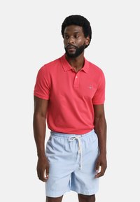 Camisa polo roja con cuello y mangas cortas, combinada con pantalones cortos de tiro ajustable en azul claro. Diseño simple y colores sólidos, material 100% algodón.