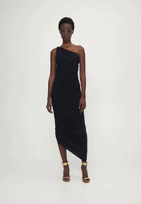 Γυναίκα που φοράει ένα μαύρο, ραμμένο φόρεμα με έναν ώμο και ασύμμετρο χείλος, χρυσά ανηρτημένα τακούνια, γυαλιά ηλίου και χρυσό κόσμημα στο λαιμό, στέκεται.