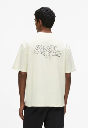 Muž otočený chrbtom, nosí krémové oversized tričko s nápisom "KARL" a štýlovou grafikou na chrbte, v kombinácii s čiernymi nohavicami.