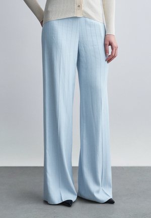 Femme portant un pantalon large bleu clair à rayures verticales et un haut côtelé beige à boutons, debout sur un sol gris devant un fond uni.
