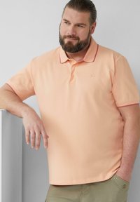 Pfirsichfarbenes Poloshirt mit kontrastierendem rotem Kragen und Ärmelbündchen, ausgestattet mit einer Zwei-Knopf-Leiste und einem kleinen Logo auf der Brust.