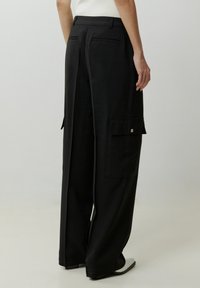 Schwarze weit geschnittene Hose mit Cargo-Taschen, glatter Textur, seitlichen Taschen und Rückendetails. Kombiniert mit einem enganliegenden Oberteil und minimalistischen Schuhen.