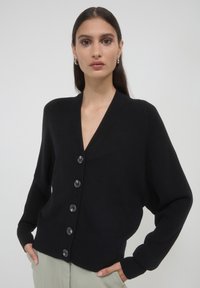 Schwarze Strickjacke mit V-Ausschnitt, sechs großen Knöpfen auf der Vorderseite und gerippten Bündchen. Der Stoff wirkt weich und leicht.