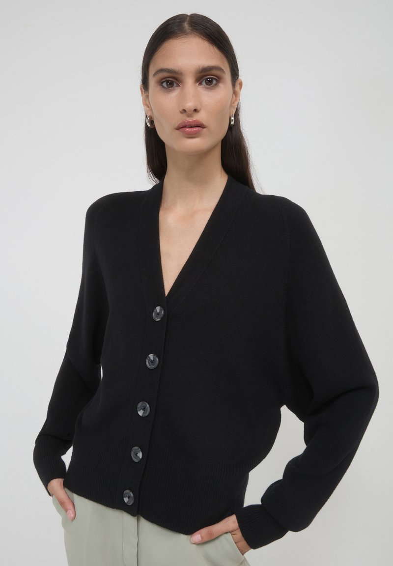 Schwarze Strickjacke mit V-Ausschnitt, sechs großen Knöpfen auf der Vorderseite und gerippten Bündchen. Der Stoff wirkt weich und leicht.