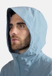 Ljusblå huvtröja i ett mjukt, vattenavvisande tyg, med synliga sömmar och dragkedjestängning fram.