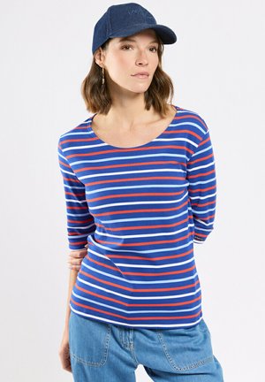 Femme portant un t-shirt à manches longues rayé bleu, rouge et blanc, un pantalon en jean bleu et une casquette de baseball bleu foncé, posant avec un bras derrière le dos.