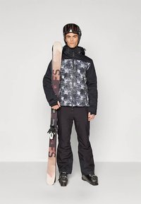 Skiausrüstung mit einer schwarz-grau getarnten Jacke und schwarzen Hosen. Die Person hält einen Ski mit einem pink-braunen Design, trägt einen schwarzen Helm und eine Schutzbrille.