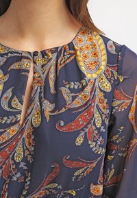 Marinblus med intrikata paisley- och blommönster i röd, gul och vit; har en rundad halsringning och knäppning.