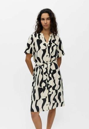 Femme aux cheveux bouclés foncés portant une robe boutonnée aux genoux, à manches courtes, avec des motifs abstraits noirs et blancs et une ceinture à la taille.