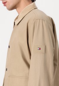 Beige jacket with a button-up design, featuring a corduroy collar and a logo patch on the sleeve. Smooth texture and slightly tailored fit.