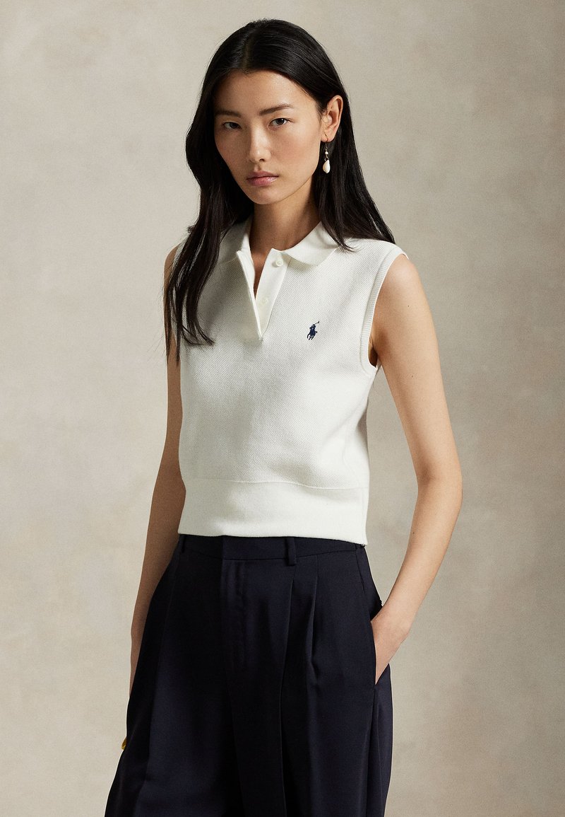 White sleeveless polo top with a textured fabric, collar, and a small blue logo on the chest, paired with dark trousers.