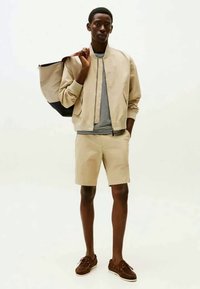 Μπέιτζ bomber τζάκετ πάνω από άσπρο-μαύρη ριγέ μπλούζα, μπέιτζ σορτς και καφέ μποέιτ παπούτσια, κρατώντας μια μαύρη και μπέιτζ τσάντα.