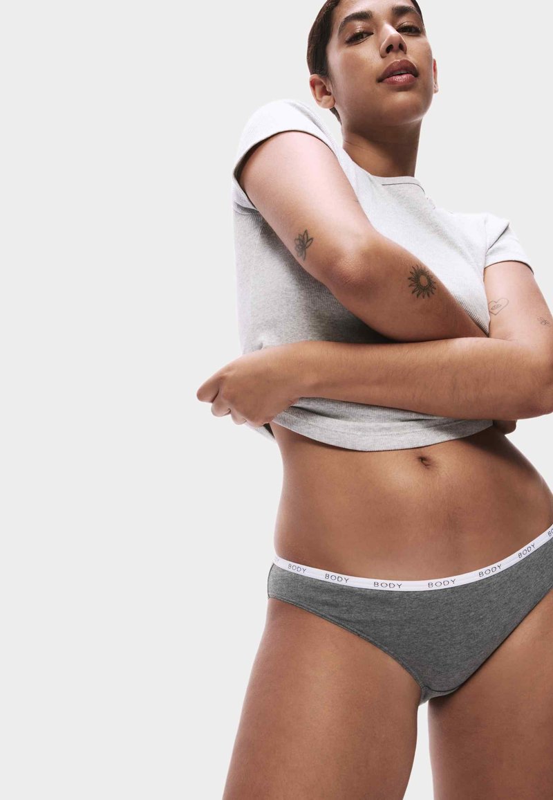 Žena v šedém spodním prádle s páskem "BODY" a krátkém světle šedém tričku, se založenýma rukama, ukazující tetování na předloktích na bílém pozadí.