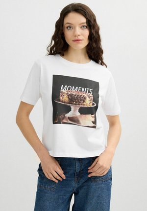 Joven con cabello castaño ondulado que lleva una camiseta blanca con un pastel de chocolate en un soporte y la palabra "MOMENTS", combinada con vaqueros azules.