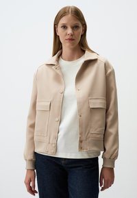 Beige jas met een kraagontwerp, twee voorzakken, drukknoopsluitingen en geribbelde manchetten; gedragen over een wit shirt en donkere jeans.