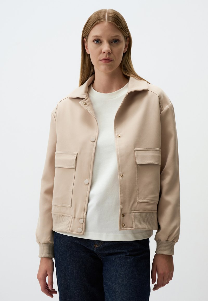 Beige jas met een kraagontwerp, twee voorzakken, drukknoopsluitingen en geribbelde manchetten; gedragen over een wit shirt en donkere jeans.