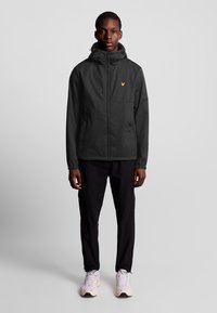Mørkegrå zip-up jakke med hætte, som har elastiske manchetter og et lille gult logo på brystet. Båret sammen med sorte bukser og lyse sneakers.