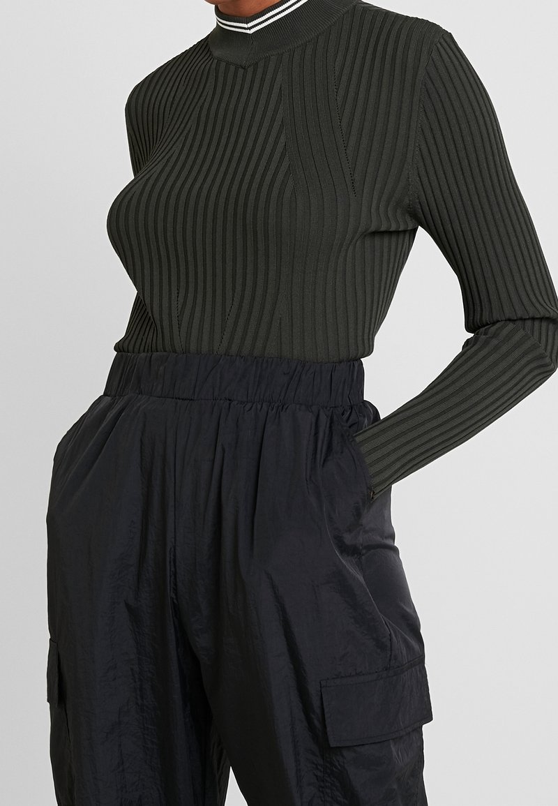 Femme portant un haut à manches longues côtelé vert foncé avec col haut et un pantalon cargo noir ample avec taille élastique et poches latérales.