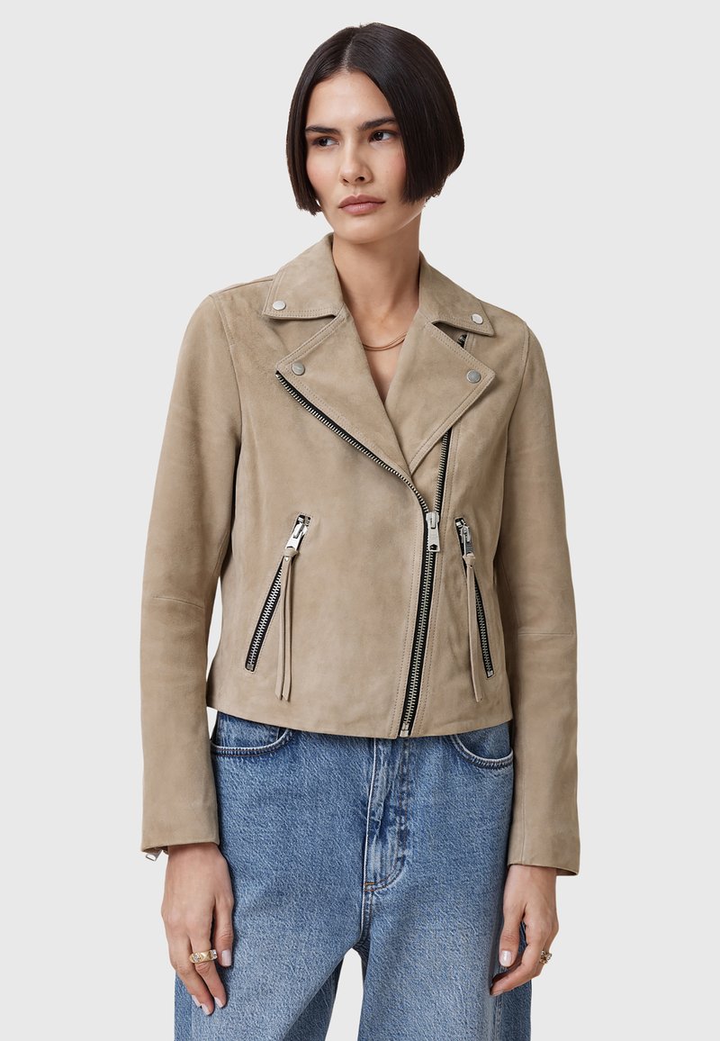 Beige ruskind bikerjakke med slagkrave, asymmetrisk lynlås og tre lynlåslommer. Har rene linjer og en tætsiddende silhuet.
