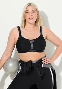 Sujeta a deportiva negra con detalles de malla y acentos grises, con tirantes anchos y una textura suave. El modelo también lleva pantalones deportivos negros y grises.