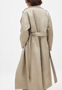 Beige pitkä trenssitakki, joka on valmistettu sileästä kankaasta. Takissa on vyöllinen vyötärö, leveät hihat ja klassinen kauluseteli.