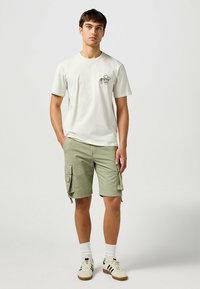 White cotton t-shirt with a black graphic on the left chest, paired with light green cargo shorts featuring side pockets and a relaxed fit.